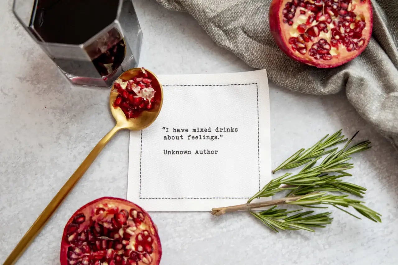White drinking quote cocktail napkin with black typewriter-style text reading, “I have mixed drinks about feelings.” – Unknown Author, styled with red wine, pomegranate, and rosemary.