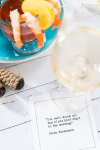 White drinking quote cocktail napkin with black typewriter-style text reading, “You can’t drink all day if you don’t start in the morning.” – Celia Rivenbark, styled beneath a wine glass with a shrimp cocktail nearby.