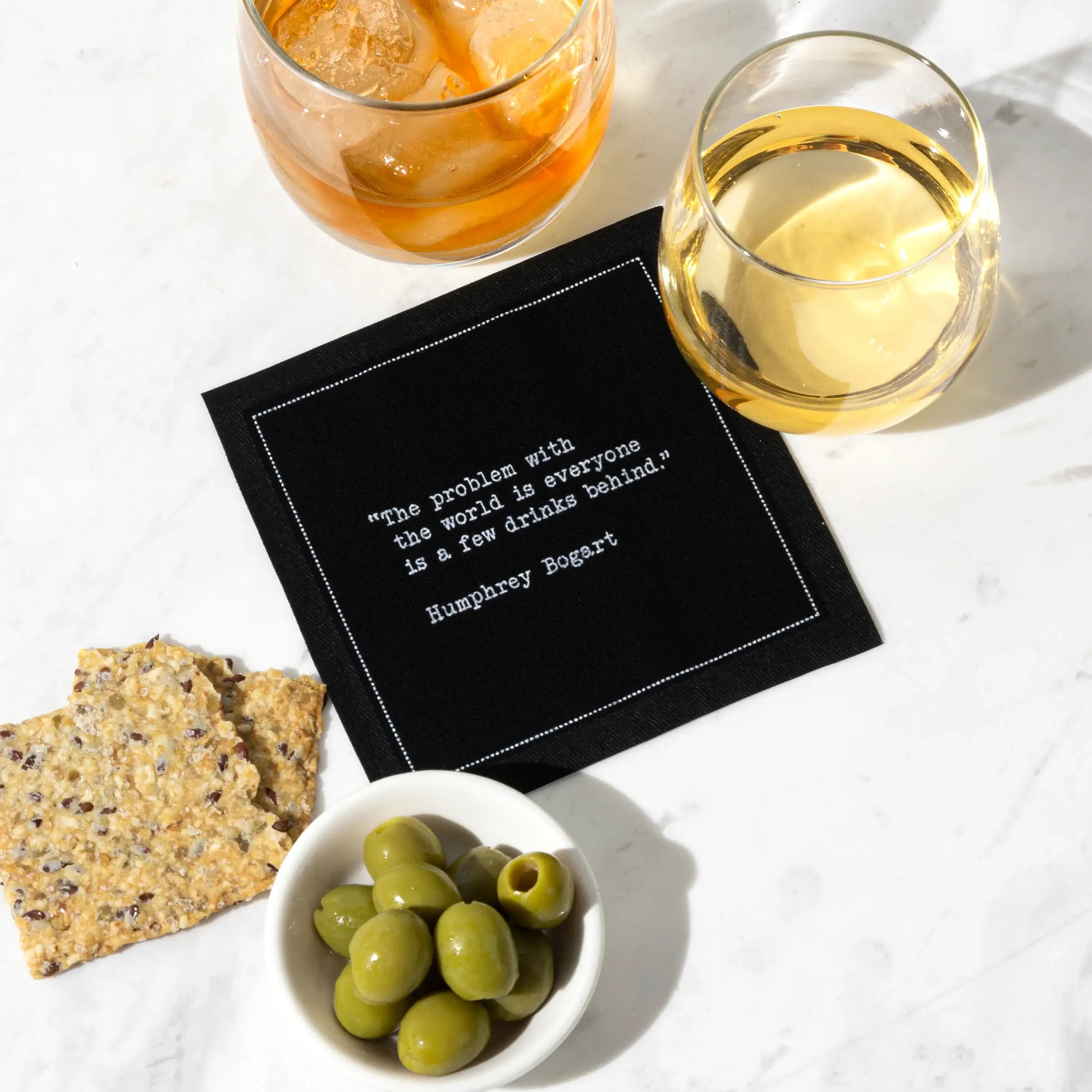 Black cocktail napkin with white typewriter-style text reading, “The problem with the world is everyone is a few drinks behind.” – Humphrey Bogart, styled with drinks, olives, and crackers.