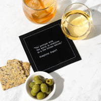Black cocktail napkin with white typewriter-style text reading, “The problem with the world is everyone is a few drinks behind.” – Humphrey Bogart, styled with drinks, olives, and crackers.