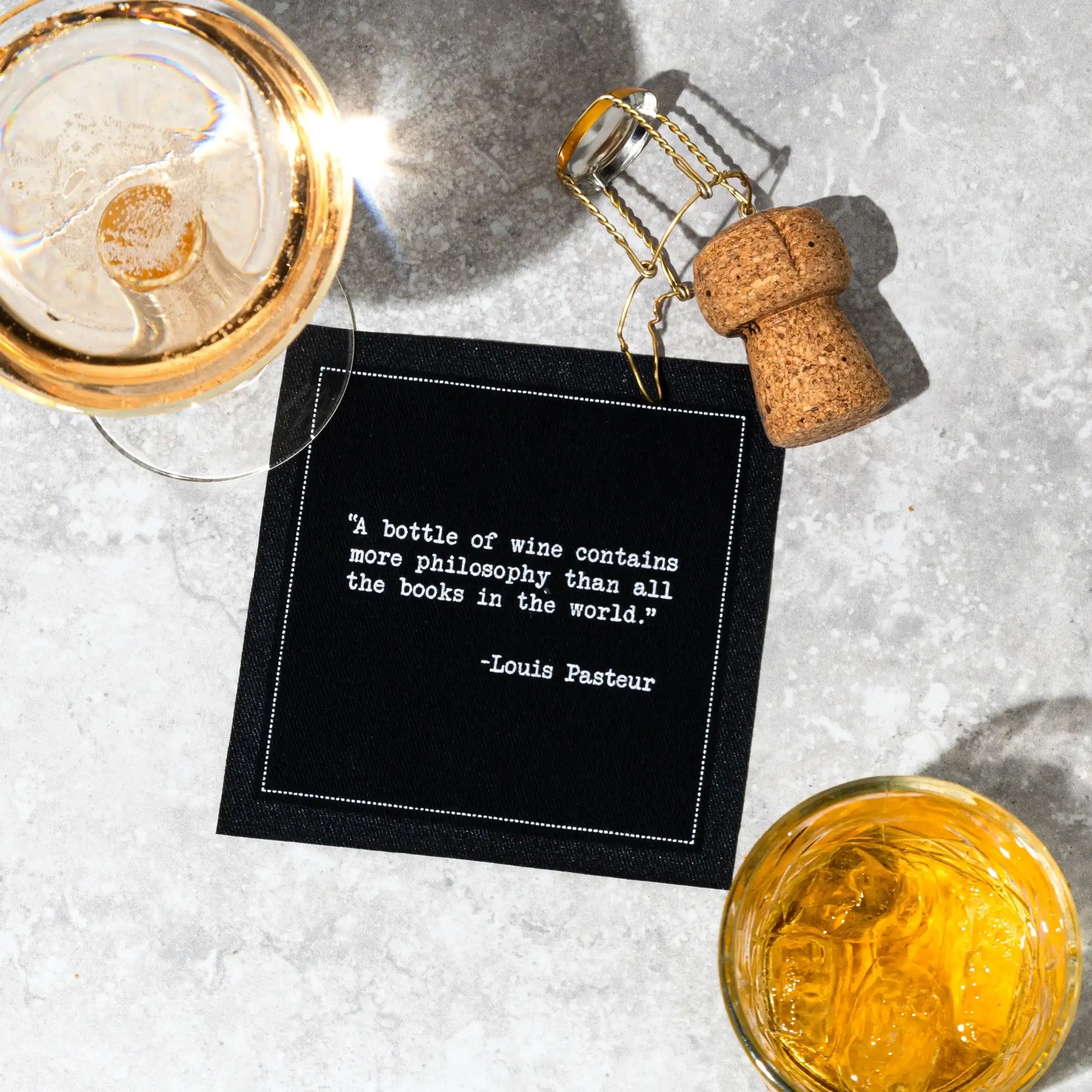 Black cocktail napkin with white typewriter-style text reading, “A bottle of wine contains more philosophy than all the books in the world.” – Louis Pasteur, styled with drinks and a champagne cork.