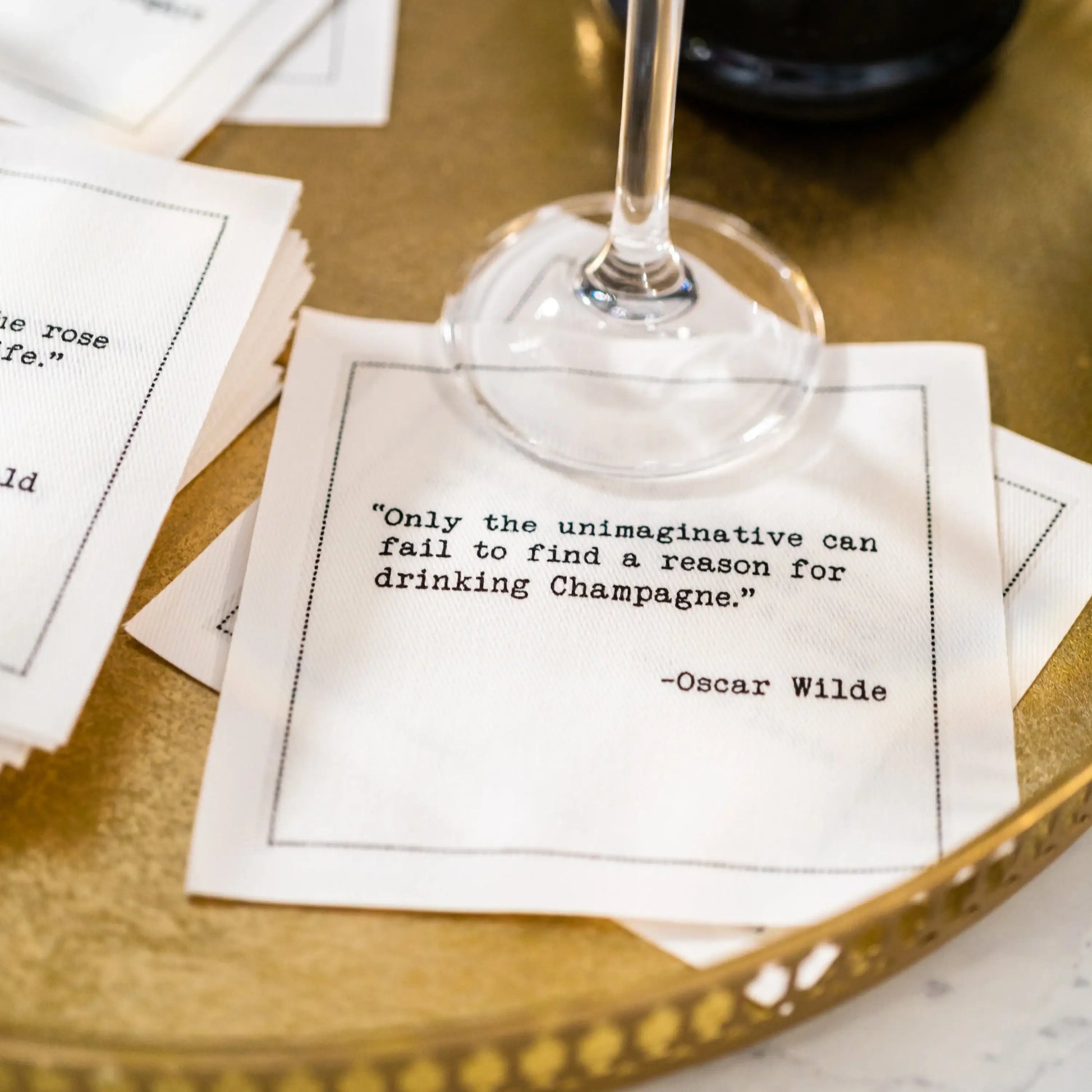 White cocktail napkin under a champagne glass featuring the quote, “Only the unimaginative can fail to find a reason for drinking Champagne.” – Oscar Wilde.