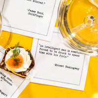 White cocktail napkin with black typewriter-style text reading, “An intelligent man is sometimes forced to be drunk to spend time with his fools.” – Ernest Hemingway, styled with drinks and an appetizer.