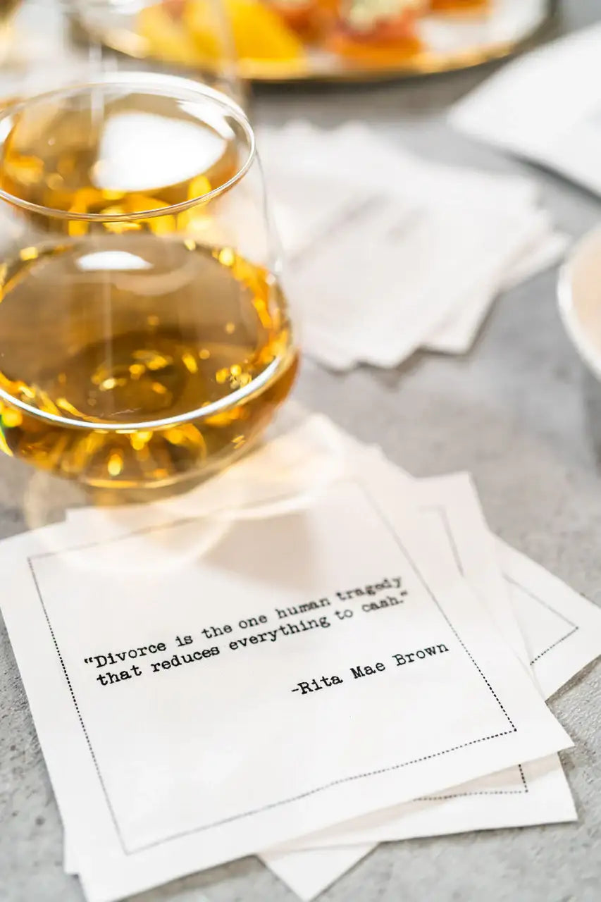 Organic cotton cocktail napkin printed with the Rita Mae Brown quote "Divorce is the one human tragedy that reduces everything to cash," styled next to a glass of whiskey on a table, from Five Star Napkins' Divorce quote collection
