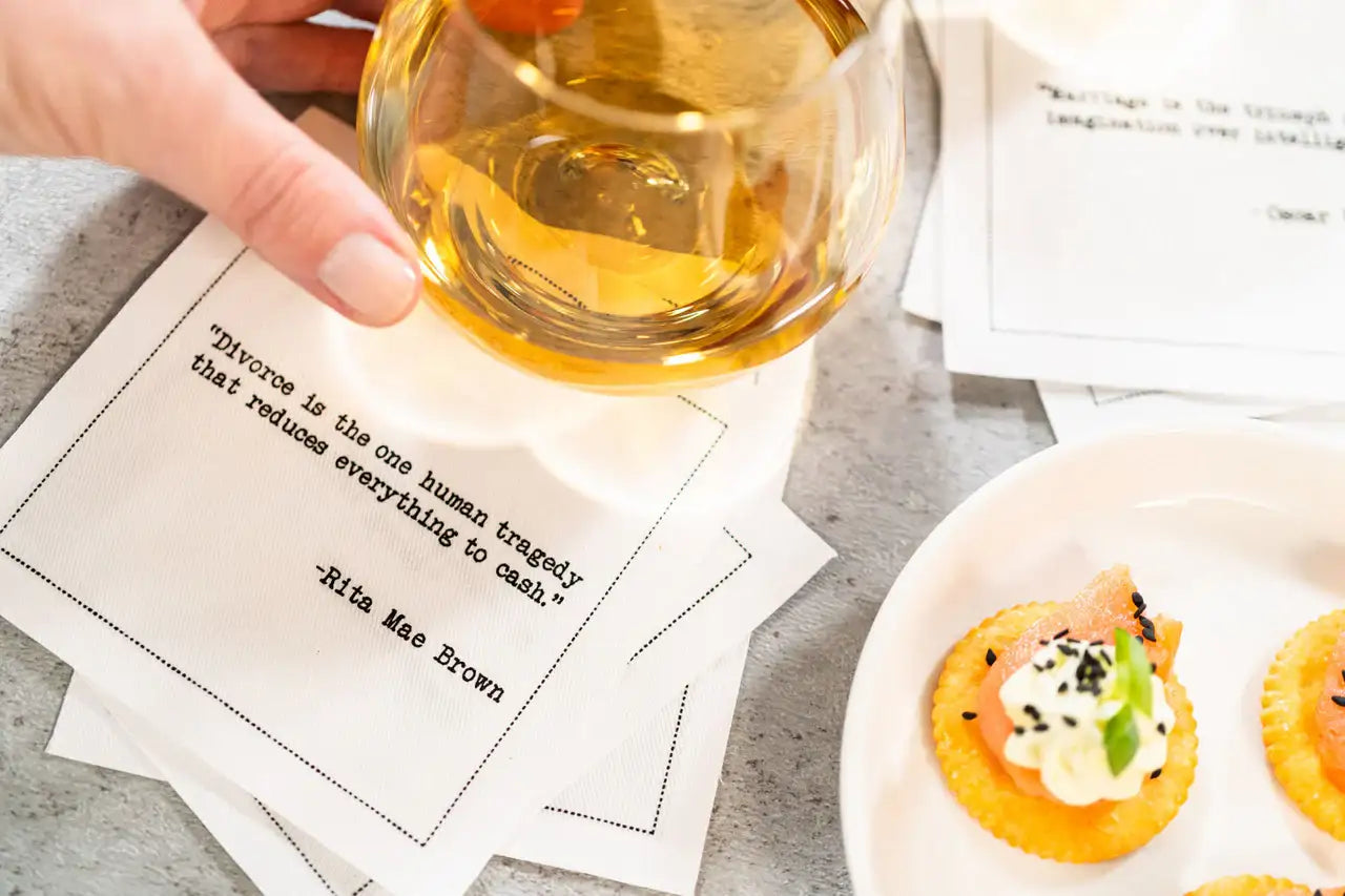 Organic cotton cocktail napkin printed with the Rita Mae Brown quote "Divorce is the one human tragedy that reduces everything to cash," styled with a hand holding a glass of whiskey and a plate of appetizers, from Five Star Napkins' Divorce quote collection
