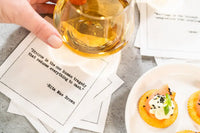 Organic cotton cocktail napkin printed with the Rita Mae Brown quote "Divorce is the one human tragedy that reduces everything to cash," styled with a hand holding a glass of whiskey and a plate of appetizers, from Five Star Napkins' Divorce quote collection