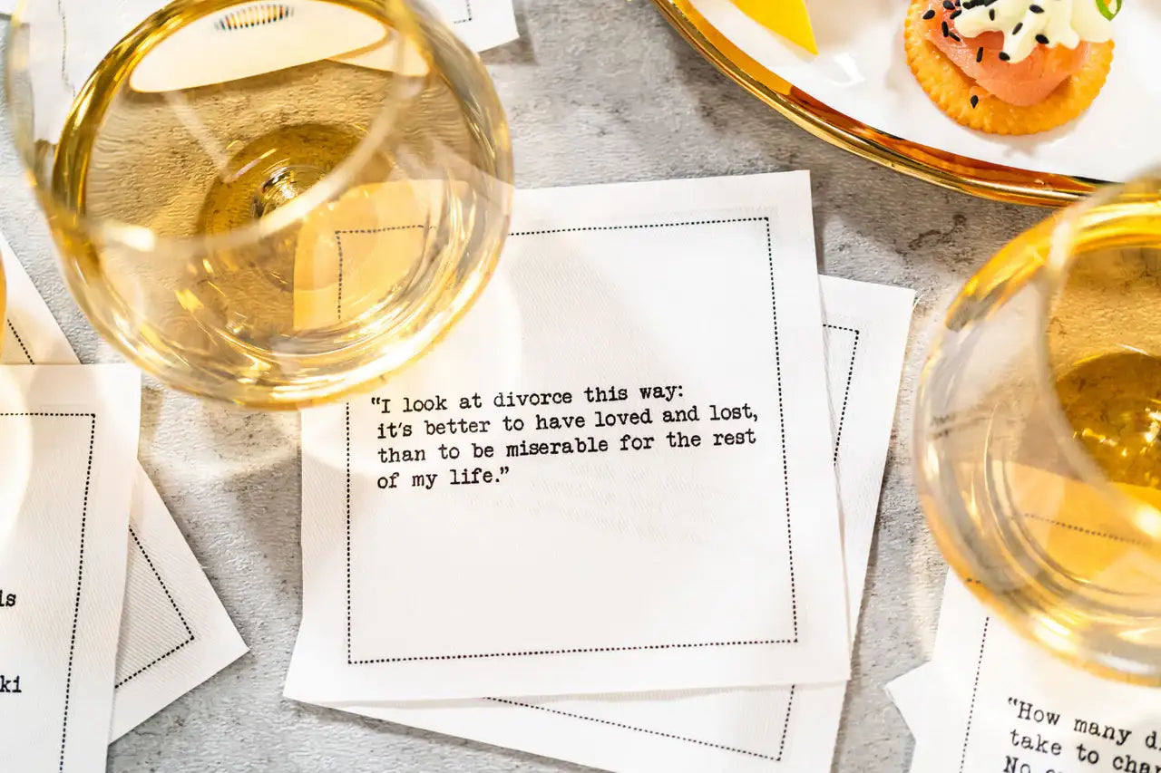 Organic cotton cocktail napkin printed with the quote "I look at divorce this way: it's better to have loved and lost, than to be miserable for the rest of my life," styled with wine glasses and appetizers on a table, from Five Star Napkins' Divorce quote collection