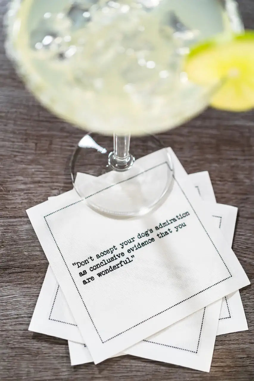 Organic cotton cocktail napkin with quote 'Don't accept your dog's admiration as conclusive evidence that you are wonderful' under a glass with ice and lime at a table setting.