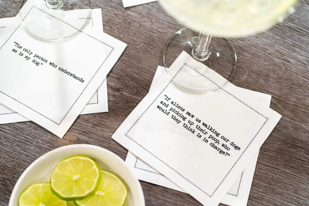 Organic cotton cocktail napkins with printed dog quotes on a wood table, drinks and lime slices nearby.