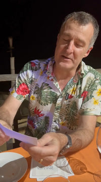 Man at a table holding an organic cotton cocktail napkin from The Drinking Quotes First Edition, with a printed quote visible in a dining setting.