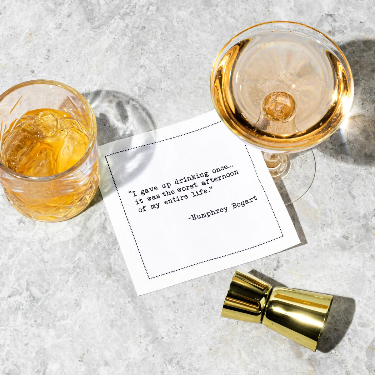White cocktail napkin with black typewriter-style text reading, “I gave up drinking once... it was the worst afternoon of my entire life.” – Humphrey Bogart, styled between two drinks.