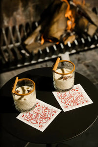 Two drinks on organic cotton napkins with red script 'Eat Drink and Be Merry!' by a fireplace.