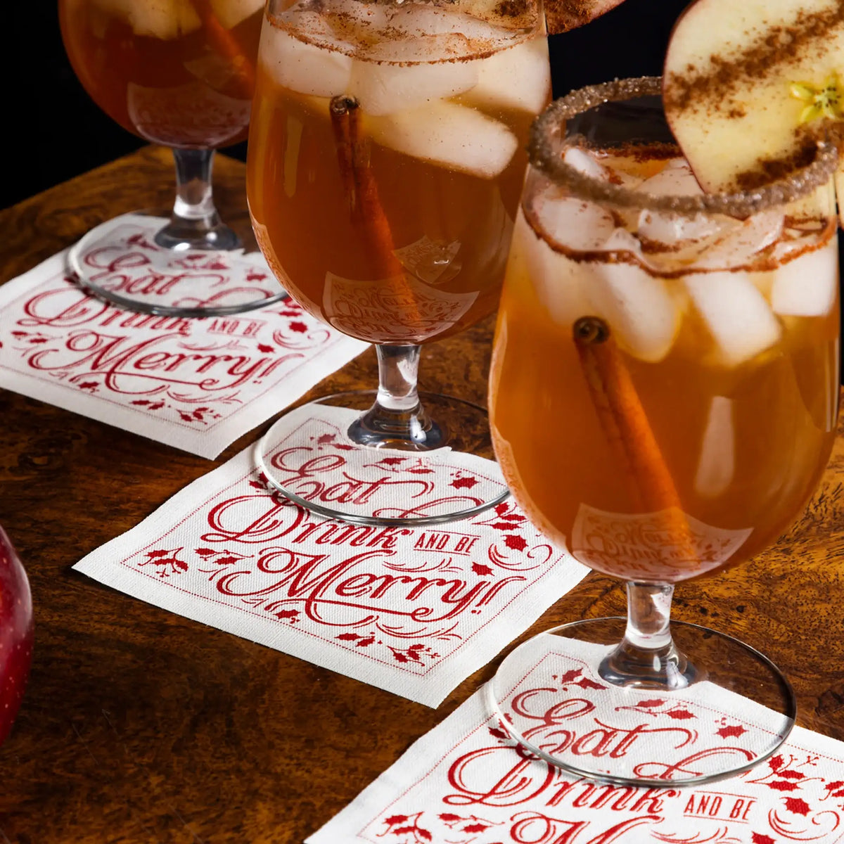 Organic cotton cocktail napkins in white with red print, quote 'Eat, Drink and be Merry!' under cocktail glasses at a table setting.