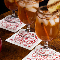 Organic cotton cocktail napkins in white with red print, quote 'Eat, Drink and be Merry!' under cocktail glasses at a table setting.