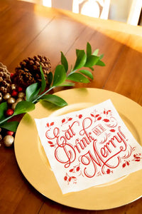 Organic cotton cocktail napkin with red 'Eat Drink and Be Merry' quote on gold plate, holiday table setting