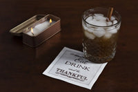Organic cotton cocktail napkin with 'Eat DRINK and be THANKFUL' quote, beside iced drink and candle on table