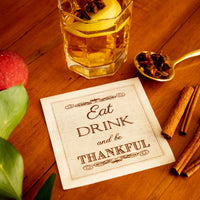 Organic cotton cocktail napkin with 'Eat DRINK and be THANKFUL' quote, styled with a drink, cinnamon sticks, and apple on a wooden table.