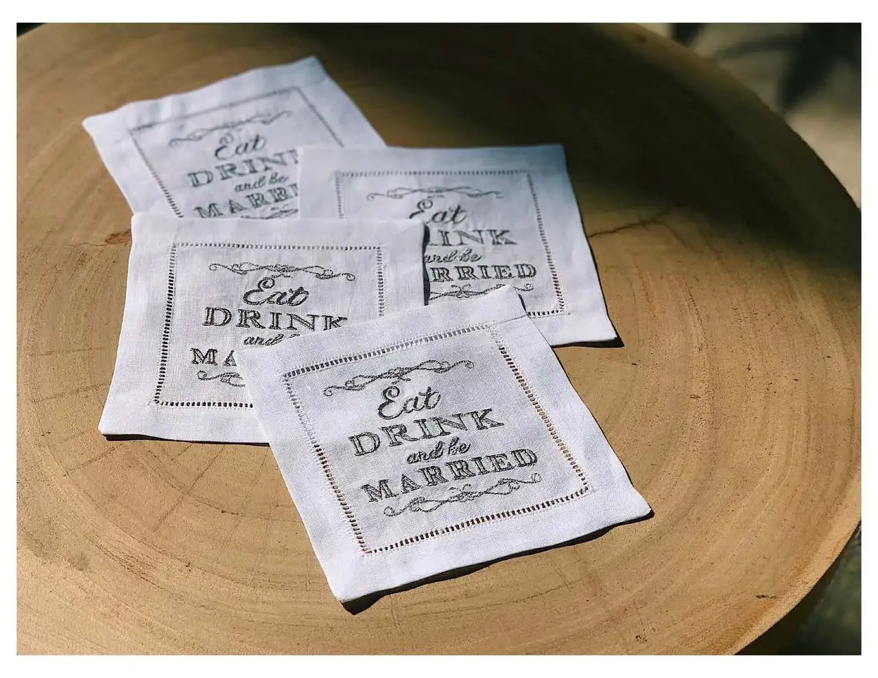 Four organic cotton cocktail napkins with 'Eat, Drink and be Married' quote on a wood table.