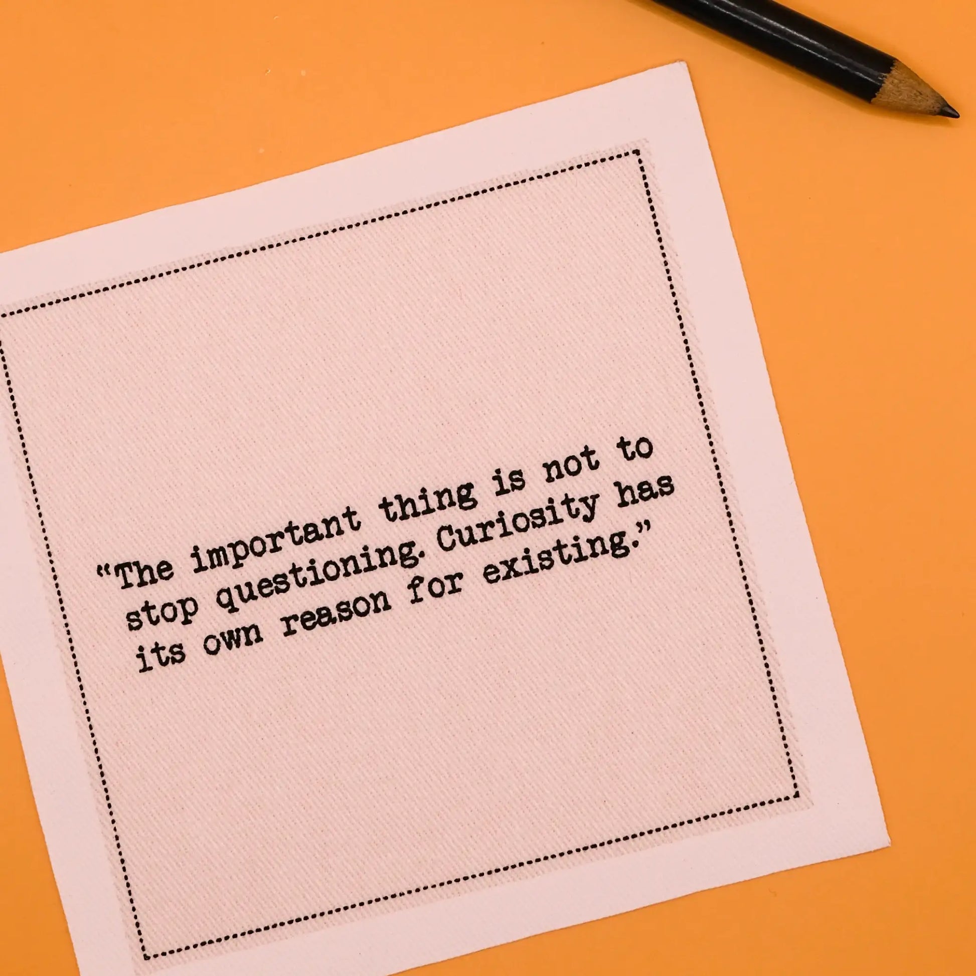 Organic cotton cocktail napkin printed with the quote "The important thing is not to stop questioning. Curiosity has its own reason for existing," styled on an orange background with a pencil, from Five Star Napkins' Einstein quote collection