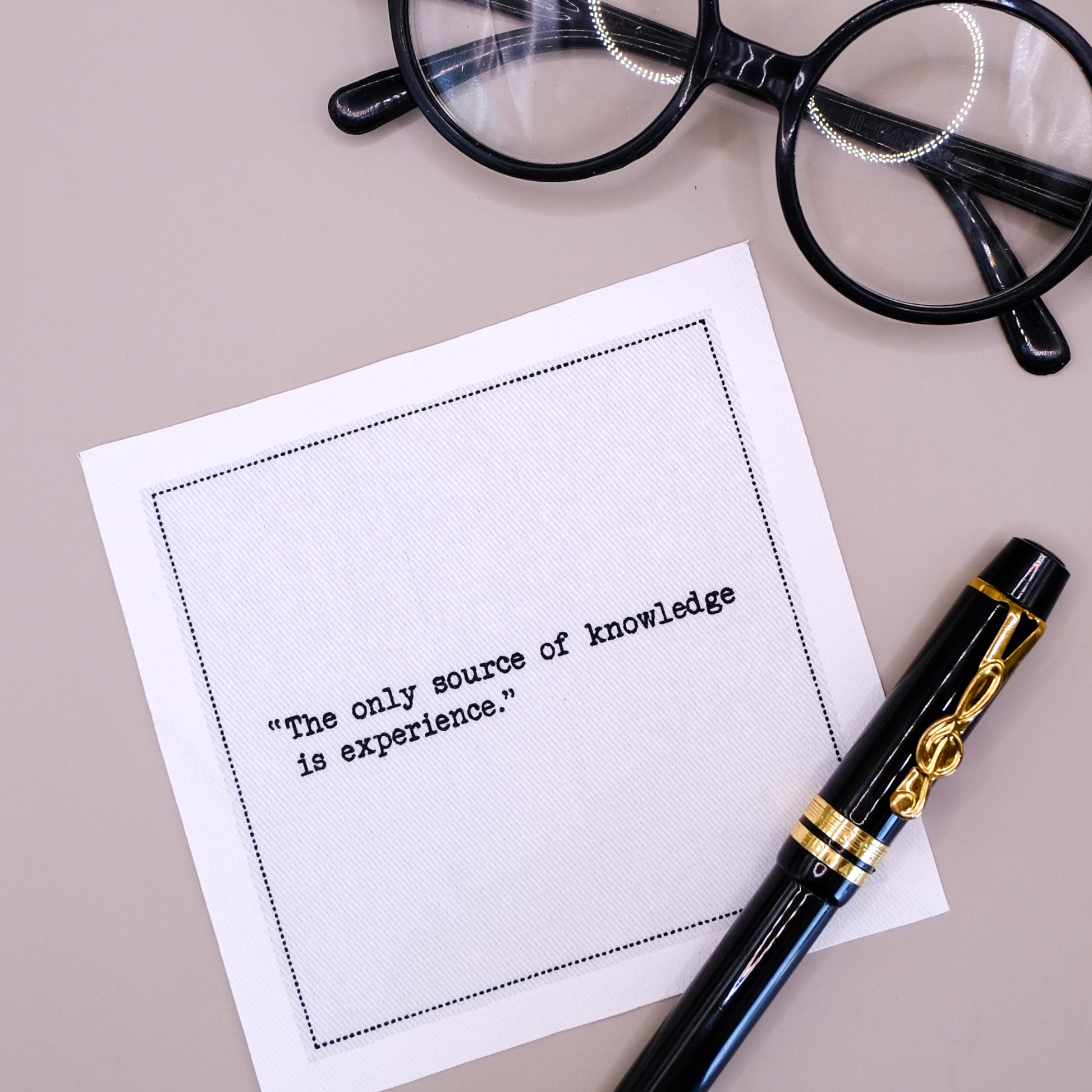 Organic cotton cocktail napkin printed with the quote "The only source of knowledge is experience," styled with a fountain pen and glasses on a tan tablecloth, from Five Star Napkins' Einstein quote collection