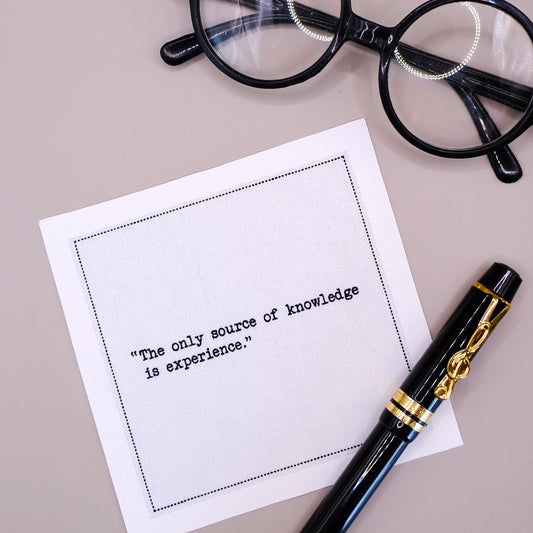 Organic cotton cocktail napkin printed with the quote "The only source of knowledge is experience," styled with a fountain pen and glasses on a tan tablecloth, from Five Star Napkins' Einstein quote collection
