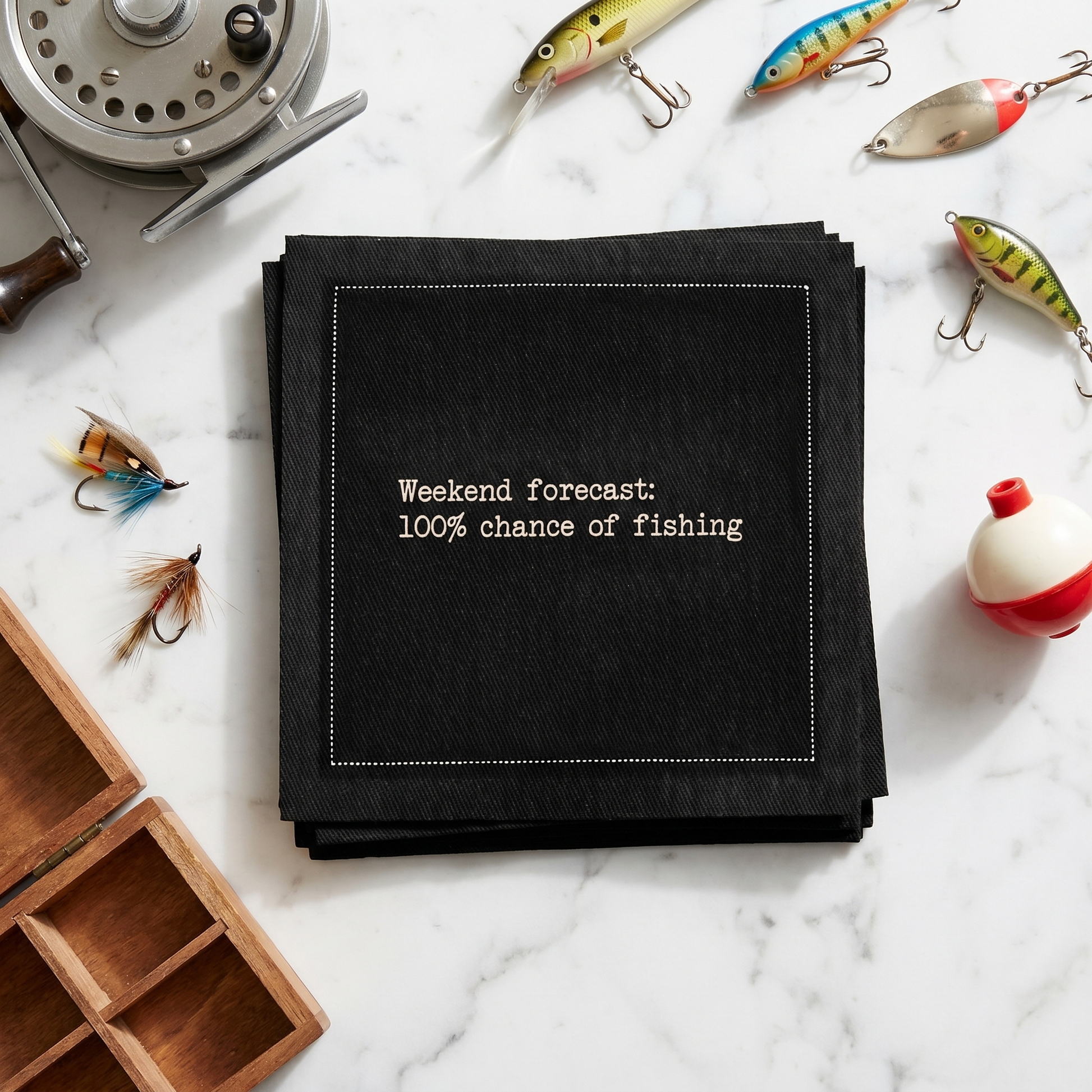 Organic cotton cocktail napkin printed with the quote "Weekend forecast: 100% chance of fishing," styled with fishing lures and tackle on a marble surface, from Five Star Napkins' Fishing Quote collection