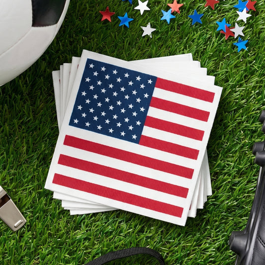 A stack of American flag cocktail napkins rests on green grass beside red, white, and blue star confetti and outdoor game equipment.