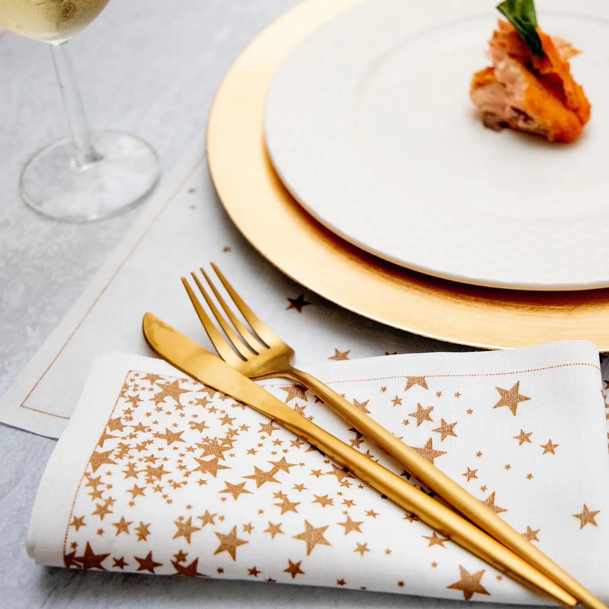 White organic cotton dinner napkin with a gold star print, folded beside gold flatware and a gold-rimmed plate, styled for a formal table setting.