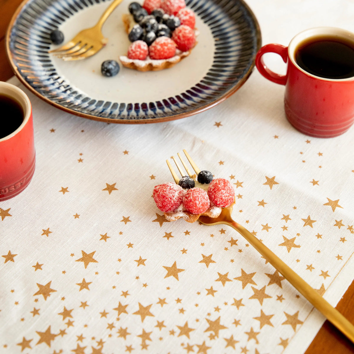 Organic cotton placemat with gold star pattern, styled for breakfast with coffee, berries, and tart.