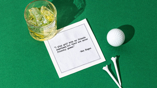 Organic cotton cocktail napkin styled with a cocktail, golf ball, and golf tees on green turf, printed with the Ben Hogan quote "I play golf with my friends sometimes, but there are never friendly games," from Five Star Napkins' Golf Legends quote collection