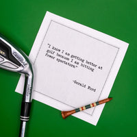 Organic cotton cocktail napkin styled with a golf club and tee on green turf, printed with the Gerald Ford quote "I know I am getting better at golf because I am hitting fewer spectators," from Five Star Napkins' Golf Humor quote collection