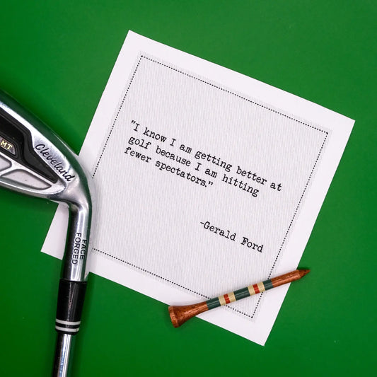 Organic cotton cocktail napkin styled with a golf club and tee on green turf, printed with the Gerald Ford quote "I know I am getting better at golf because I am hitting fewer spectators," from Five Star Napkins' Golf Humor quote collection