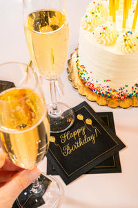 Two hands toasting with champagne beside a birthday cake and black organic cotton cocktail napkins printed with “Happy Birthday!” in gold script, by Five Star Napkins