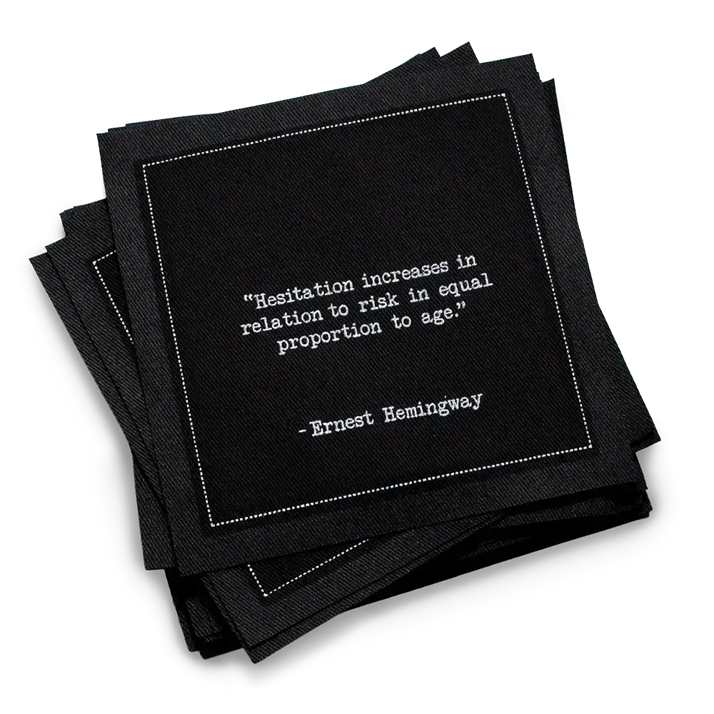Organic cotton cocktail napkin, black, with white dotted border printed with the quote "Hesitation increases in relation to risk in equal proportion to age," from Five Star Napkins' Hemingway quote collection
