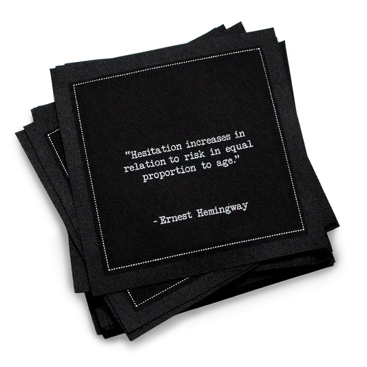 Organic cotton cocktail napkin, black, with white dotted border printed with the quote "Hesitation increases in relation to risk in equal proportion to age," from Five Star Napkins' Hemingway quote collection