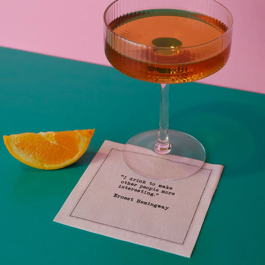 Organic cotton cocktail napkin printed with the quote "I drink to make other people more interesting," styled with a cocktail glass and an orange slice on a pink and teal background, from Five Star Napkins' Hemingway quote collection