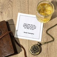 Organic cotton cocktail napkin printed with the quote "Only those who are prepared to go too far can possibly know how far they can go," styled on a wood table with a glass of whiskey, a leather journal, and a pocket watch, from Five Star Napkins' Hemingway quote collection