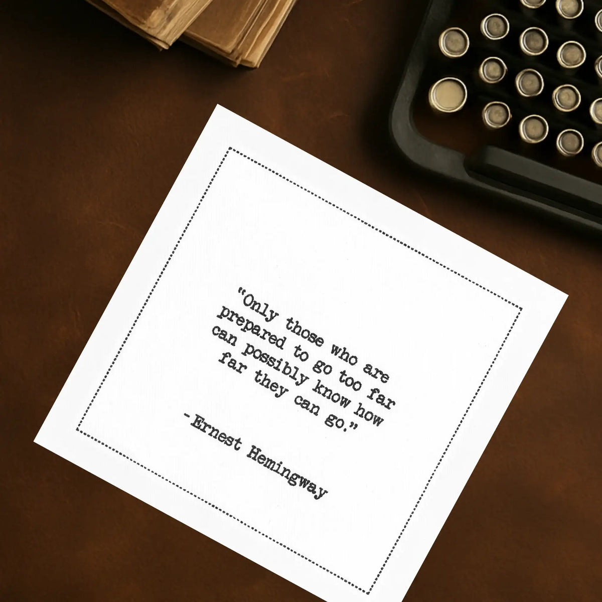 Organic cotton cocktail napkin printed with the quote "Only those who are prepared to go too far can possibly know how far they can go," styled next to a vintage typewriter and stack of old books, from Five Star Napkins' Hemingway quote collection