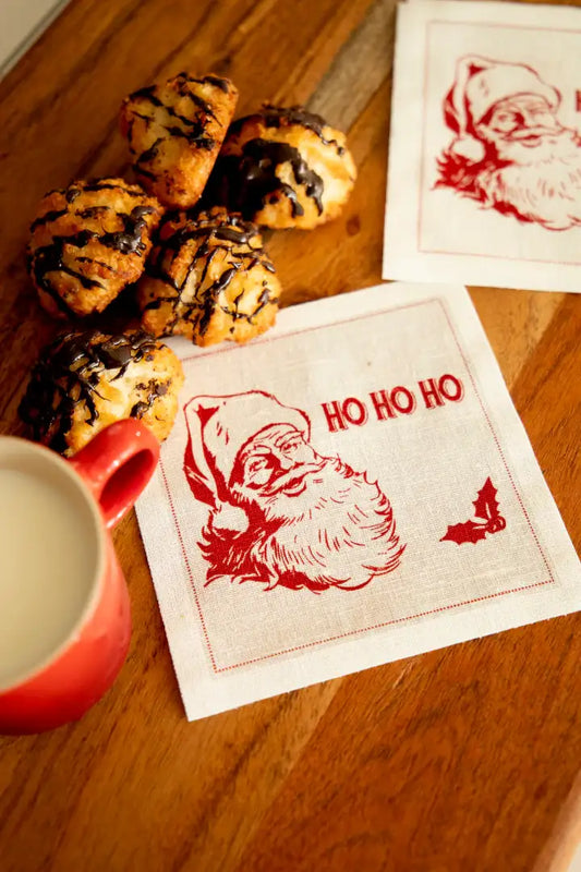 Organic cotton cocktail napkin with red Santa and 'HO HO HO' quote, cookies, and milk on table