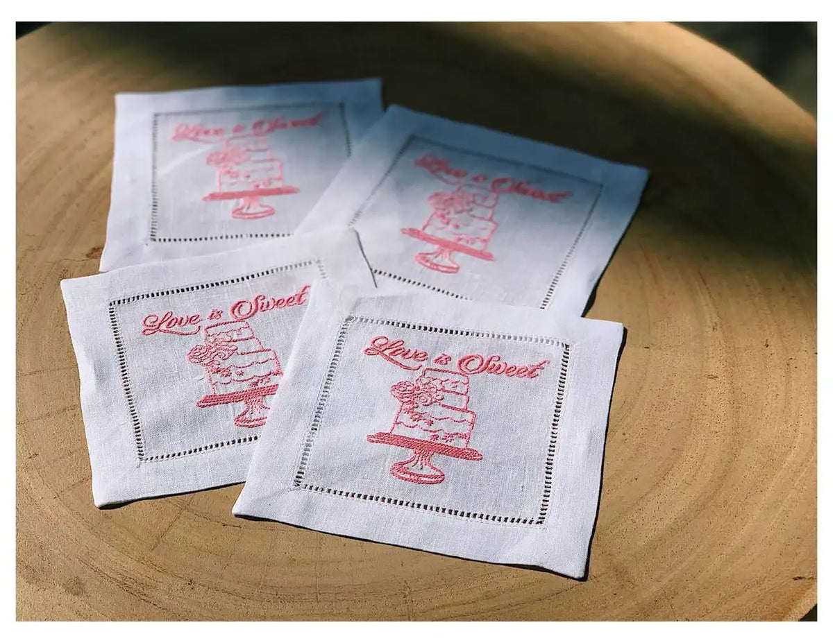 Four organic cotton cocktail napkins with pink 'Love is Sweet' quote and cake illustration on a wooden table