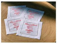 Four organic cotton cocktail napkins with pink 'Love is Sweet' quote and cake illustration on a wooden table