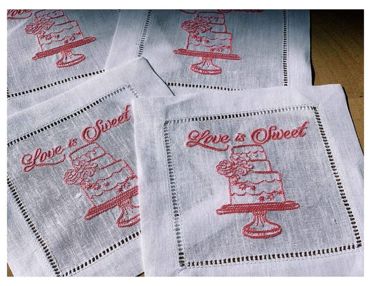 Organic cotton napkins with pink embroidered cake and 'Love is Sweet' quote, styled flat.