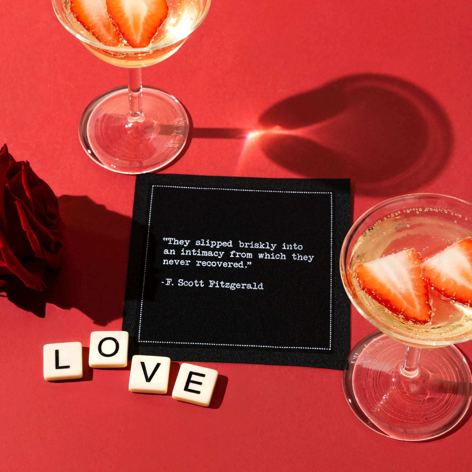 A black Love Quotes cocktail napkin on a red surface reads, “They slipped briskly into an intimacy from which they never recovered.” —F. Scott Fitzgerald. Strawberry drinks, red roses, and letter tiles spelling “LOVE” surround it.