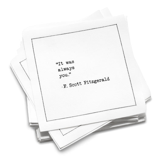 A stack of white Love Quotes cocktail napkins with black dotted borders. The top napkin reads, “It was always you.” —F. Scott Fitzgerald.