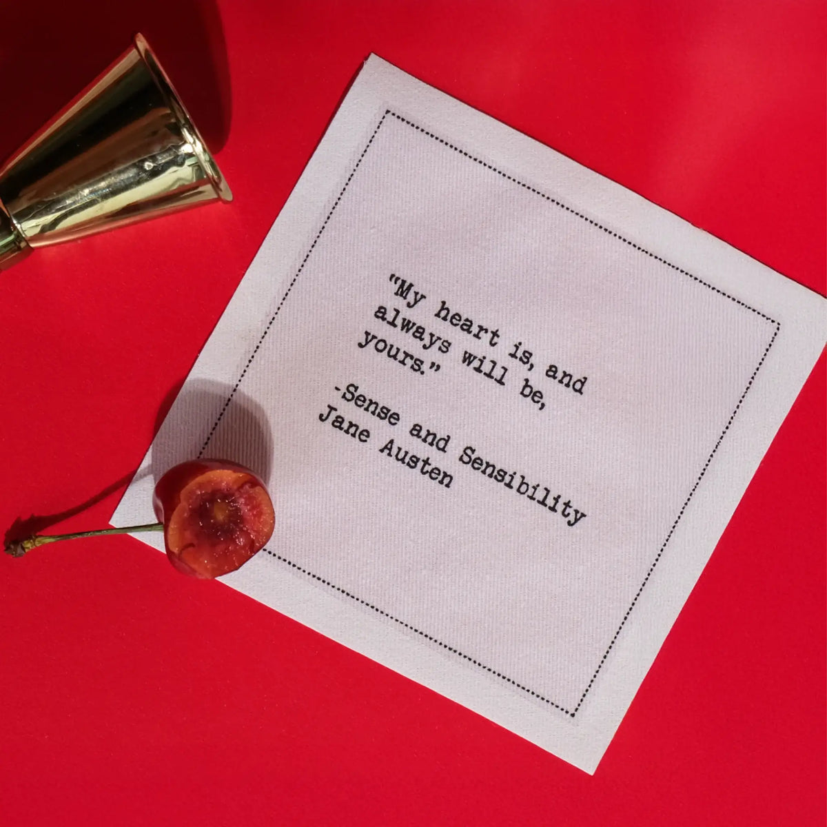 A white Love Quotes cocktail napkin on a bright red surface, accented with a cherry and silver jigger. The napkin reads, “My heart is, and always will be, yours.” —Jane Austen, Sense and Sensibility.