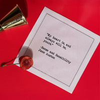 A white Love Quotes cocktail napkin on a bright red surface, accented with a cherry and silver jigger. The napkin reads, “My heart is, and always will be, yours.” —Jane Austen, Sense and Sensibility.