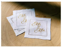 Three organic cotton cocktail napkins with gold 'Mr. & Mrs.' embroidery on wood surface