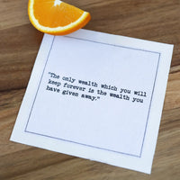 Organic cotton cocktail napkin printed with the quote "The only wealth which you will keep forever is the wealth you have given away," styled on a wood table with an orange slice, from Five Star Napkins' Marcus Aurelius quote collection