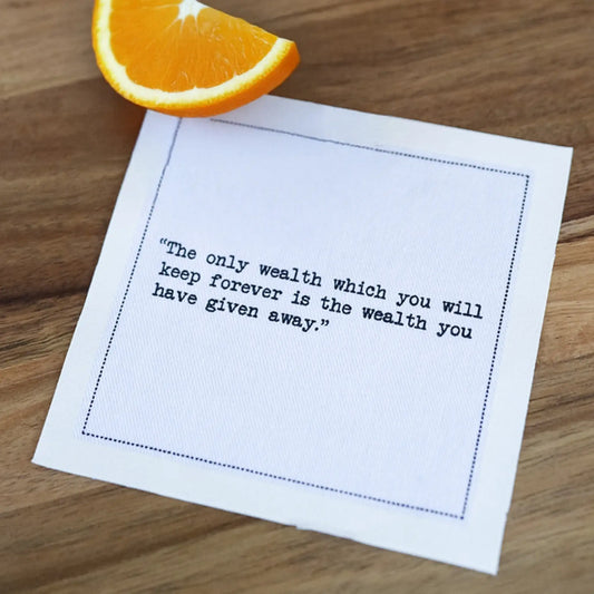 Organic cotton cocktail napkin printed with the quote "The only wealth which you will keep forever is the wealth you have given away," styled on a wood table with an orange slice, from Five Star Napkins' Marcus Aurelius quote collection