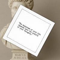 Organic cotton cocktail napkin printed with the quote "The happiness of your life depends upon the quality of your thoughts," styled in front of a marble bust of Marcus Aurelius, from Five Star Napkins' Marcus Aurelius quote collection